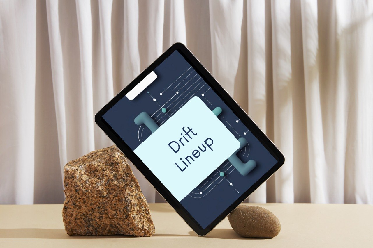 A tablet device propped open on two stones against a soft beige curtain backdrop. The screen displays a dark blue interface with a light blue card reading 'Drift Lineup'.