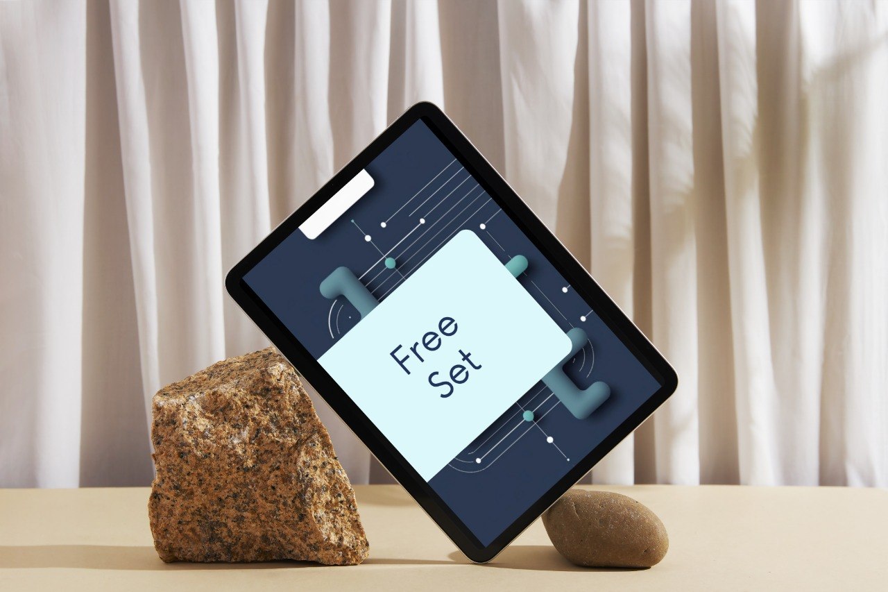 A tablet device shown at an angle on a tabletop, leaning against a small stone, with a large light blue card reading 'Free Set' on its screen. The background features draped cream curtains.